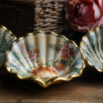 A large scallop shell, with a decoupage design of floral patterns. The shell is gold gilded. It is staged on a beautiful background